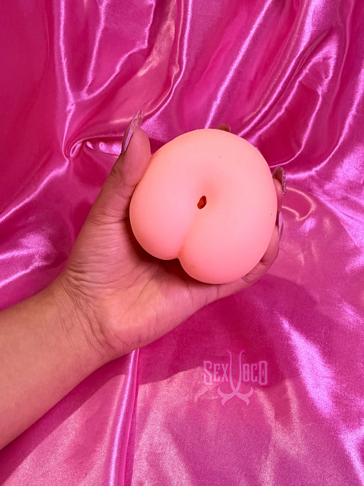 Masturbador pequeño en forma de corazón color piel siendo sostenido por manos con manicura sobre fondo rosa Sex Shop Xoco.
