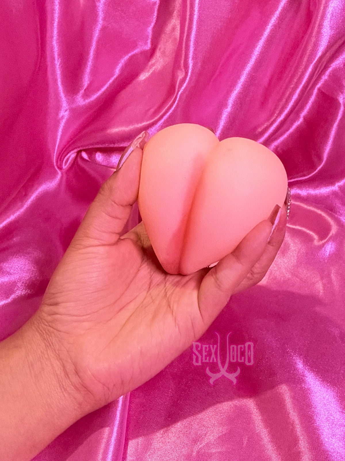 Masturbador pequeño en forma de corazón color piel siendo sostenido por manos con manicura sobre fondo rosa Sex Shop Xoco.