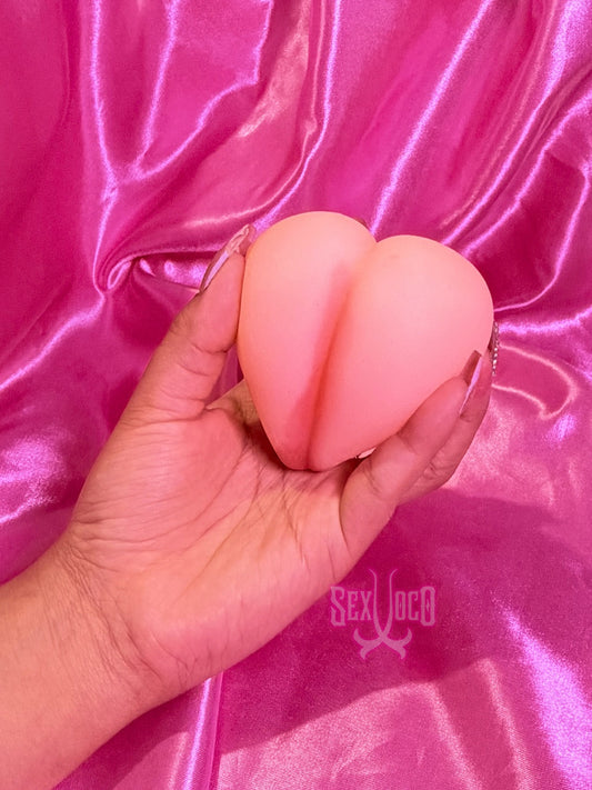 Masturbador pequeño en forma de corazón color piel siendo sostenido por manos con manicura sobre fondo rosa Sex Shop Xoco.